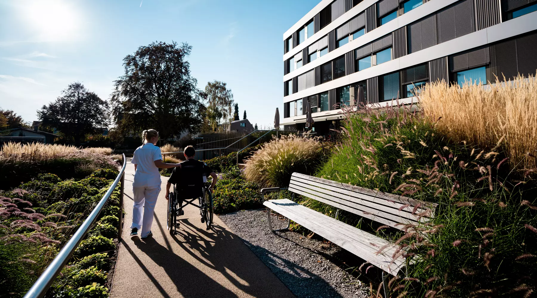 Vue extérieure de l'HFR Meyriez-Murten, un patient en chaise roulante accompagné par une infirmière se promène en fin d'après-midi au soleil
