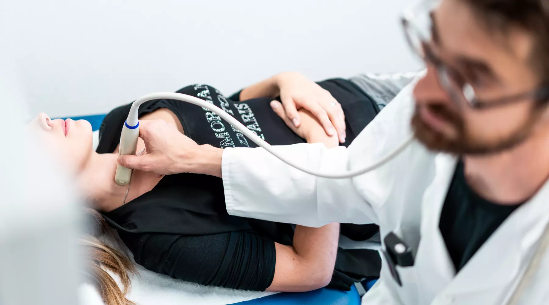 Photo d'un examen: une jeune femme, allongée sur un lit tandis que le médecin effectue une échographie au niveau de se gorge.