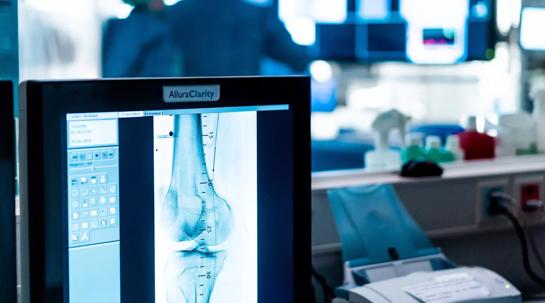 A l'avant-plan, un écran affiche le résultat d'un examen d'imagerie médicale, à l'arrière-plan, séparé par une vitre, deux médecins effectuent une intervention