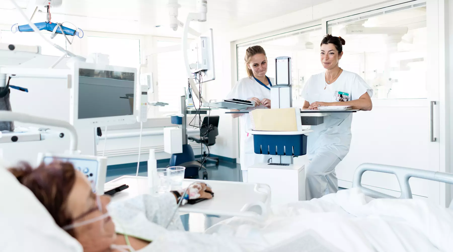 Vue d'une chambre des soins intensifs. Le personnel médical-soignant, au chevet de la patiente, consulte des dossiers