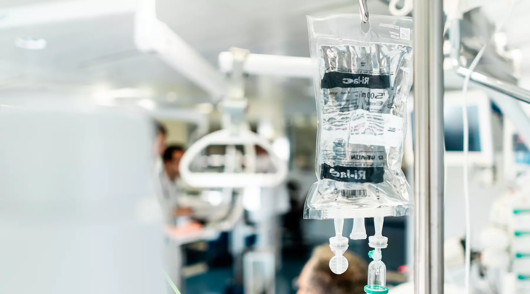 Gros plan sur une poche de perfusion à l'avant plan. A l'arrière plan on distingue un patient allongé dans un lit.