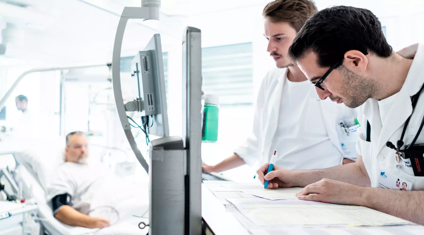 Deux médecins étudient les données sur un écran d'ordinateur. Un patient est visible à l'arrière-plan.