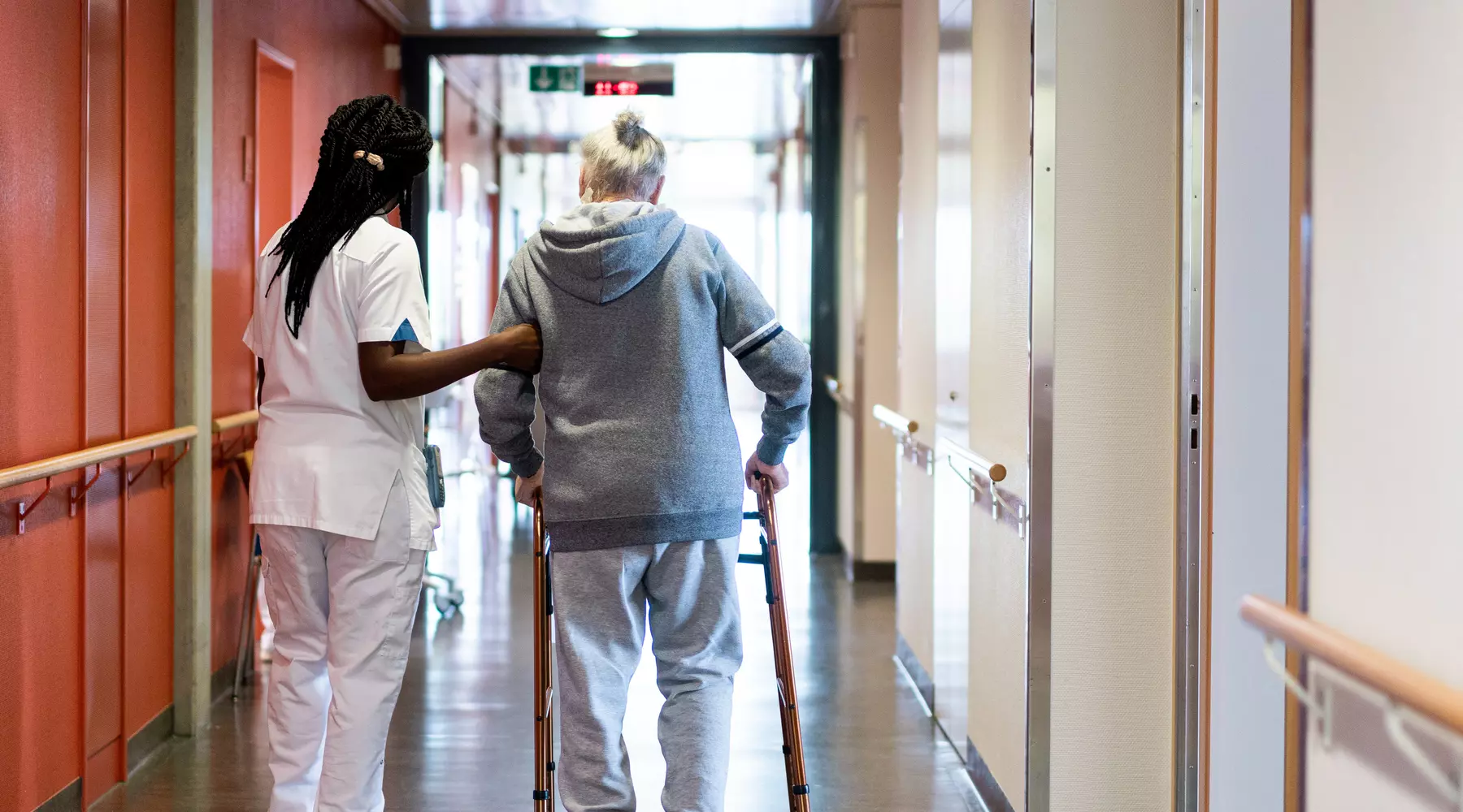 Photo d'un couloir d'hôpital. Une infirmière soutien une personne en train de marcher avec un déambulateur. Les deux personnes sont vues de dos. 