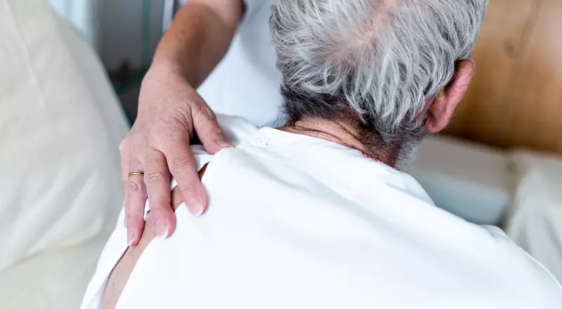 Gros plan sur la main d'une infirmière, délicatement posée sur le dos d'un patient âgé. 