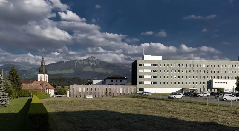 Aussenansicht des HFR Riaz im Sommer 2018 von der Seite des Haupteingangs mit dem Parkplatz. Im Hintergrund sind Berge sowie die Kirche von Riaz zu sehen. 